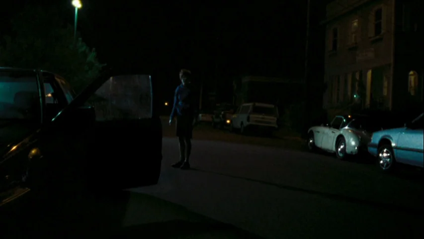Wendy and Lucy (2008) movie still: wide — A quiet residential street at night with parked cars and a distant building. A lone figure stands near an…