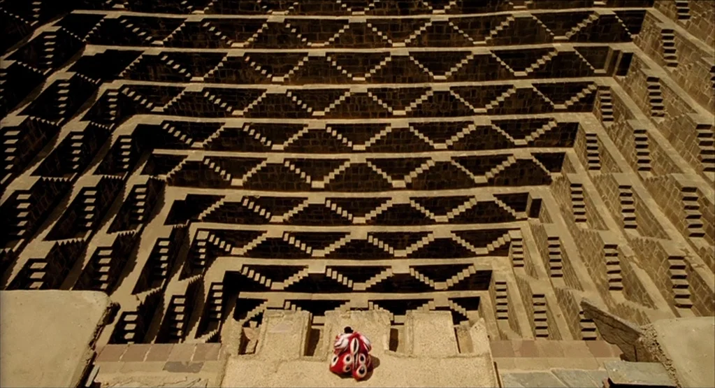 The Fall movie still: wide — An ancient stone stepwell features a complex, descending pattern of symmetrical carved staircases. A lone figure in…