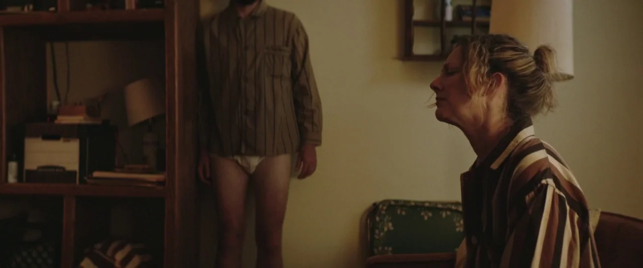 Lemon (2017) movie still: medium — A dimly lit room features a wooden shelving unit and an armchair. A man in pajamas stands by the wall while a…