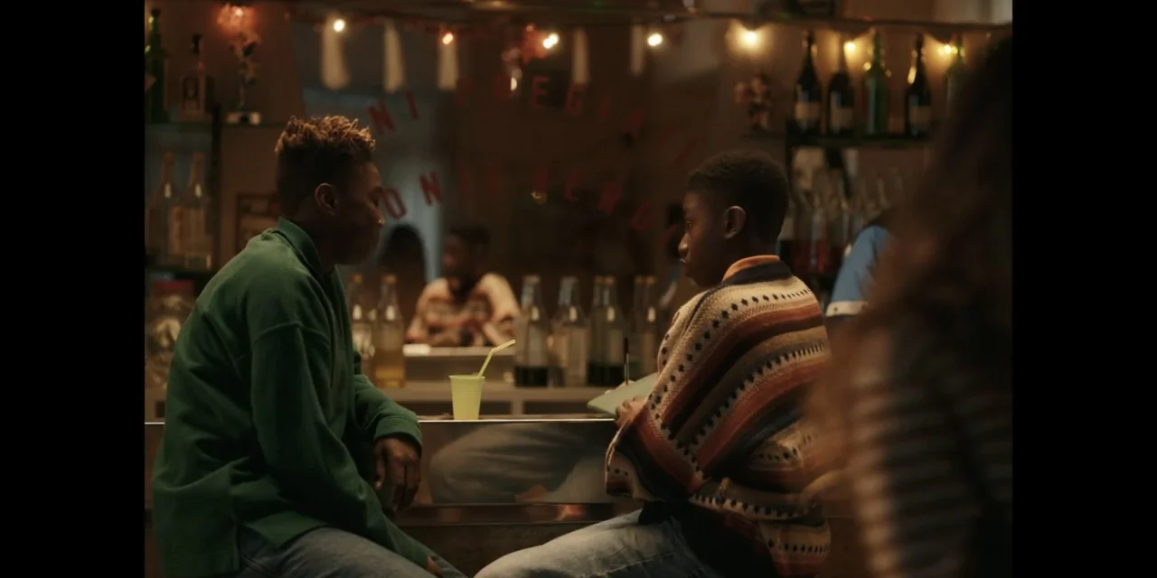 Autumn Beat (2022) movie still: medium — Two individuals sit at a dimly lit bar with bottles on shelves behind them. The two men talk to each other…