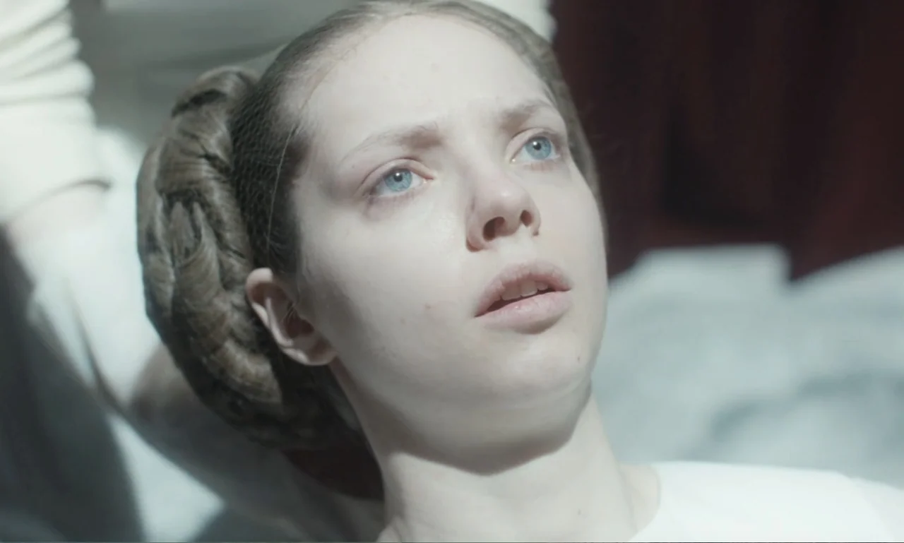 The Ugly Stepsister (2025) movie still: closeup — A bright interior space features a person positioned in the foreground. The individual gazes…
