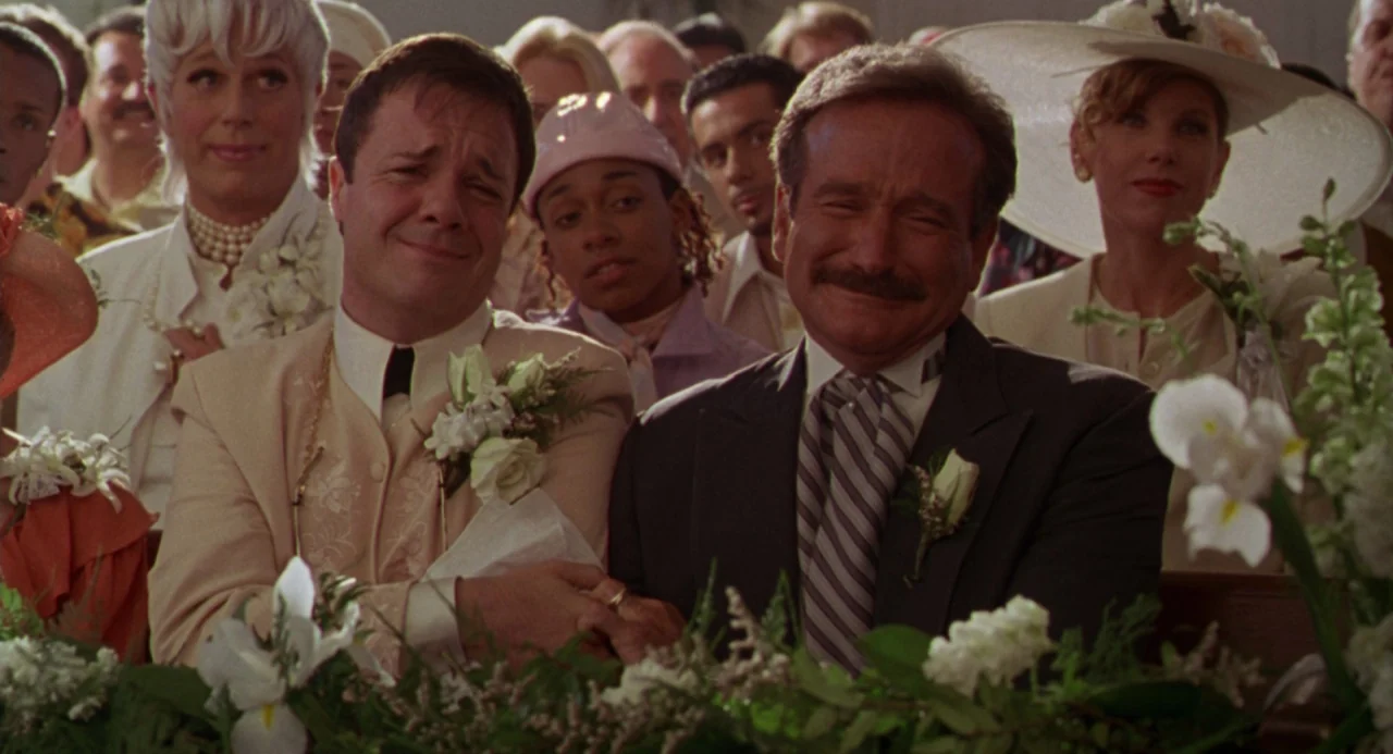 The Birdcage (1996) movie still: medium — A crowded church pews setting filled with wedding guests in formal attire. Two men sit together in the…