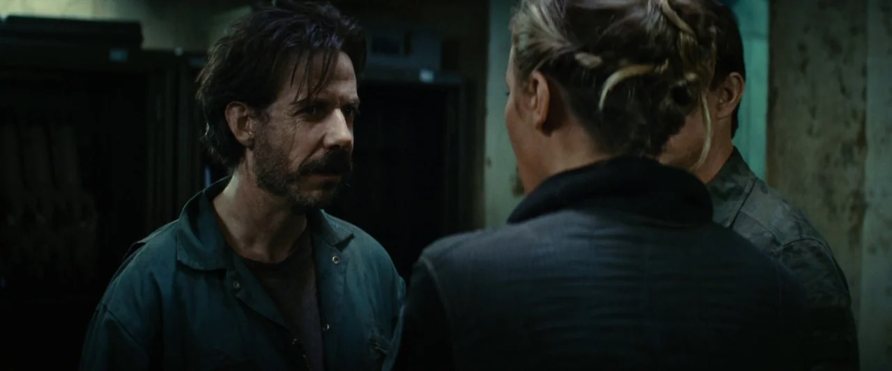 Edge of Tomorrow (2014) movie still: medium — A dimly lit industrial room features metal storage lockers and textured concrete walls. A man in a…