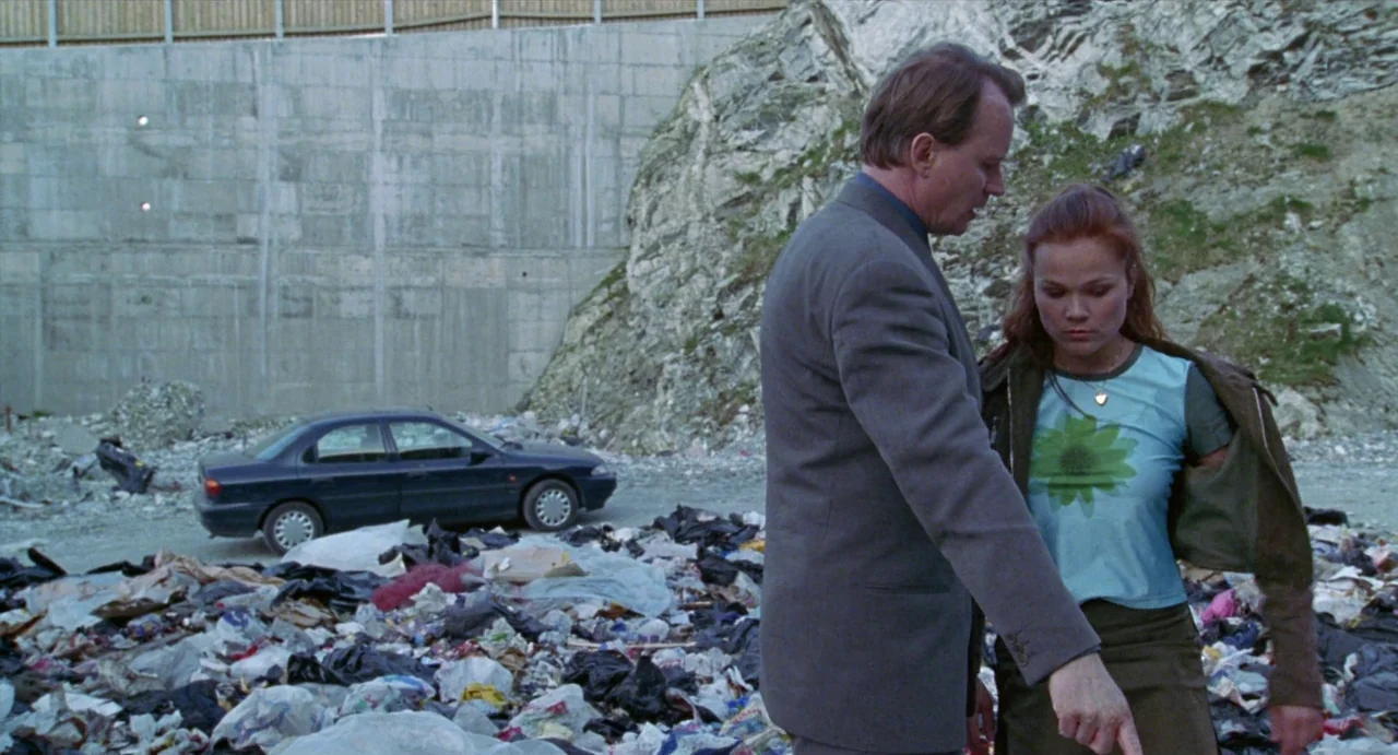 Insomnia (1997) movie still: medium — A man and woman stand in a littered landfill against a high concrete barrier. The man points toward the ground…