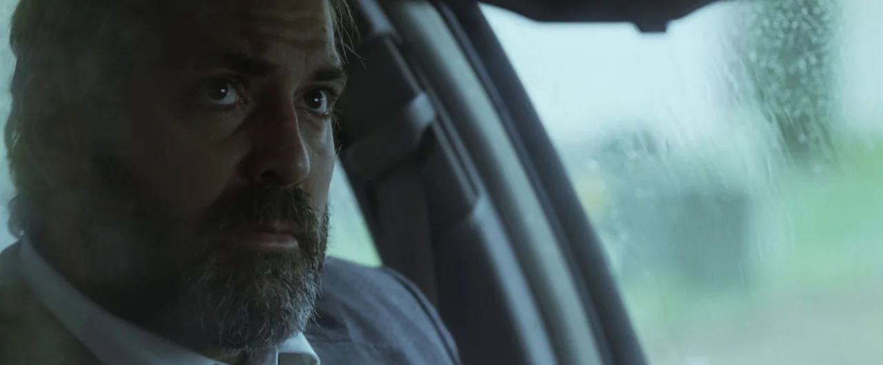 Syriana (2005) movie still: closeup — The interior of a car during a rainy day with water droplets on the glass. A middle-aged man with a full beard…