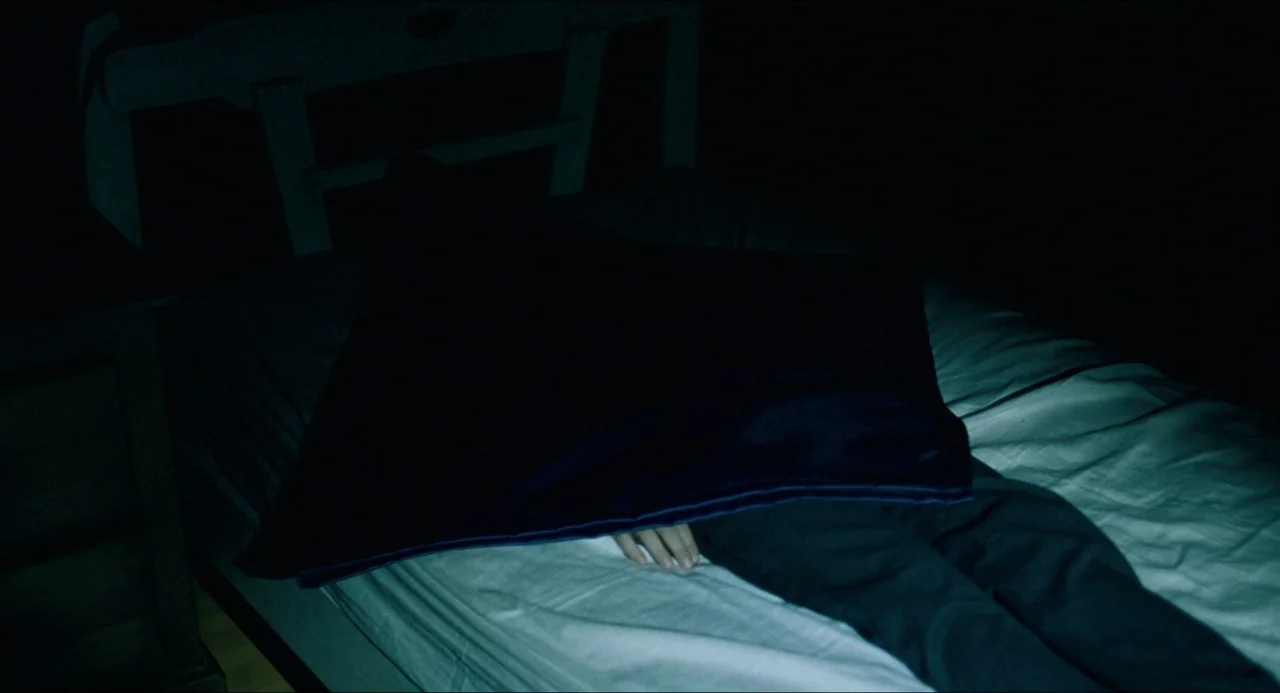 The Lodge (2019) movie still: medium — A dark bedroom shows a person lying flat on a bed. They are completely covered by a dark blanket with only…