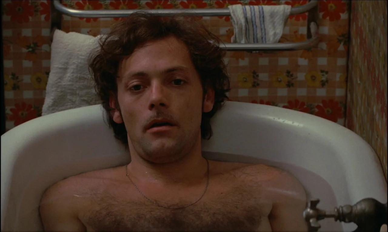 Serie Noire (1979) movie still: closeup — The background consists of patterned floral wallpaper with a metal towel rack above. A shirtless man with…