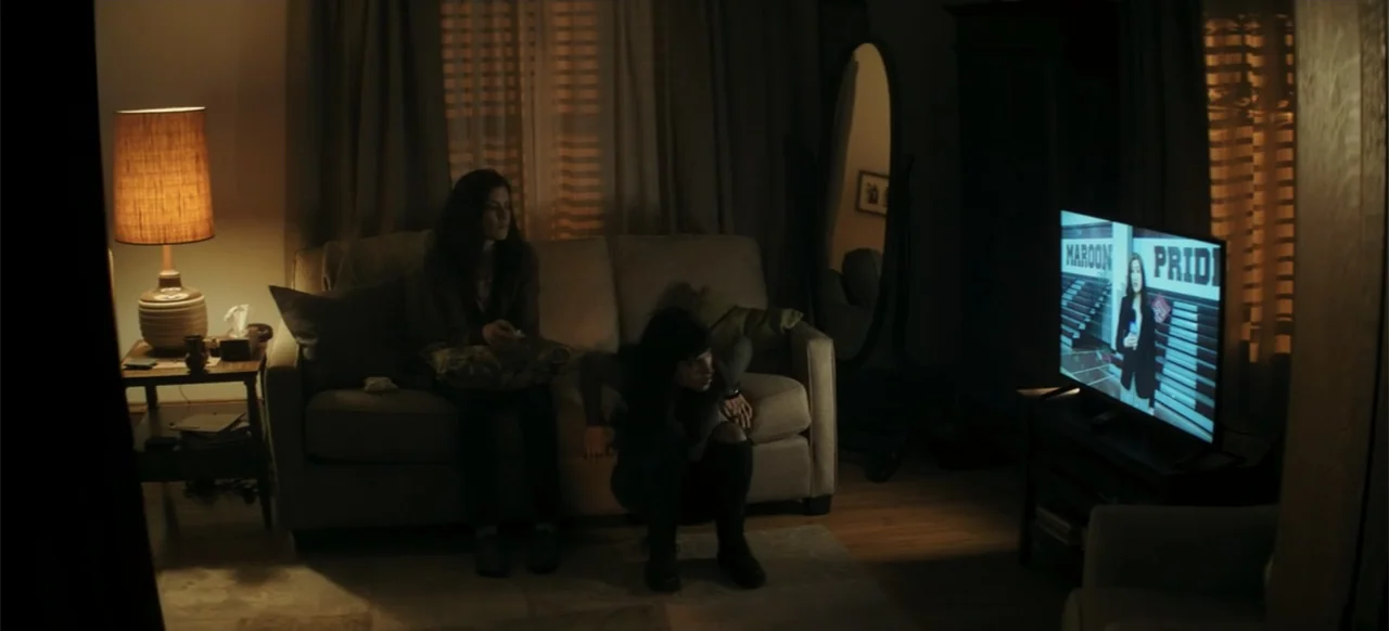 I Still See You (2018) movie still: medium — A dimly lit living room contains a sofa, a floor lamp, and a television. Two women watch a television…