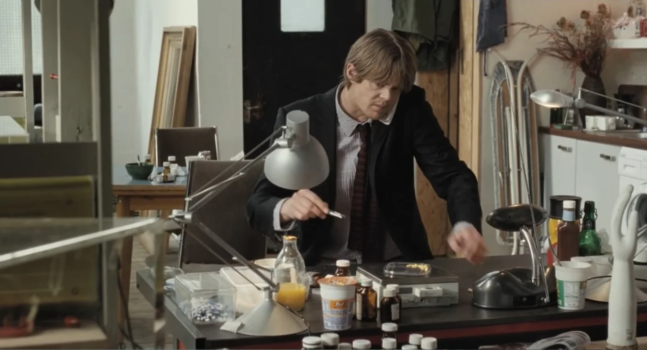 Death at a Funeral (2010) movie still: medium — A cluttered home office contains a desk filled with numerous medicine bottles and equipment. A man…