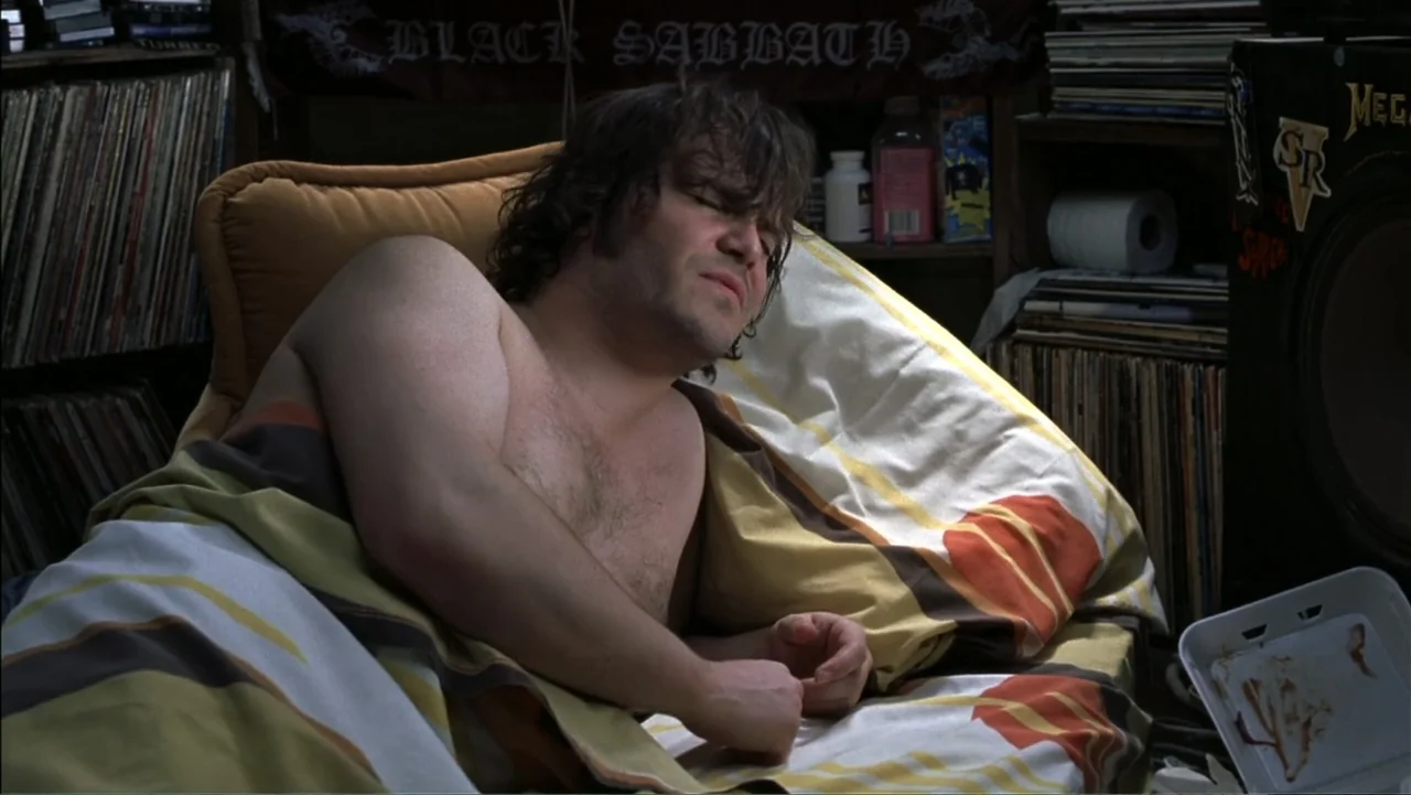School of Rock (2003) movie still: medium — A cluttered bedroom filled with stacks of vinyl records and personal items. A man lies in bed covered by…