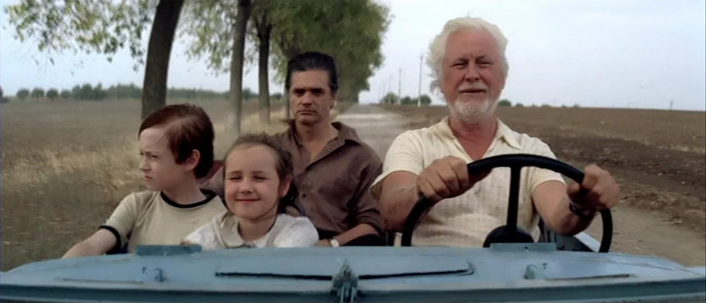 The Banishment (2007) movie still: medium — A group of people sits in an open-topped vehicle on a rural dirt road. A line of trees borders one side…