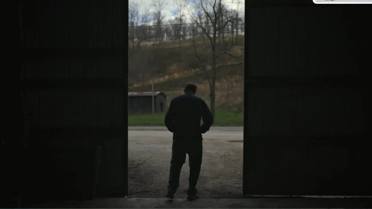 Murder at Carter County Mines (2023) movie still: fullbody — A dark, empty barn interior opens to a gravel path and rural hillside landscape. A man…