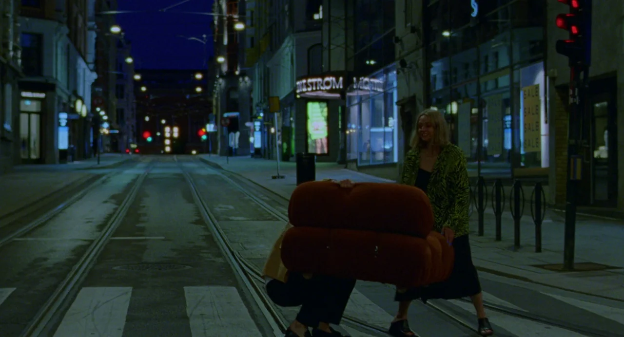 Sick of Myself (2022) movie still: fullbody — An empty city street at night features tram tracks and storefronts with bright signage. Two…