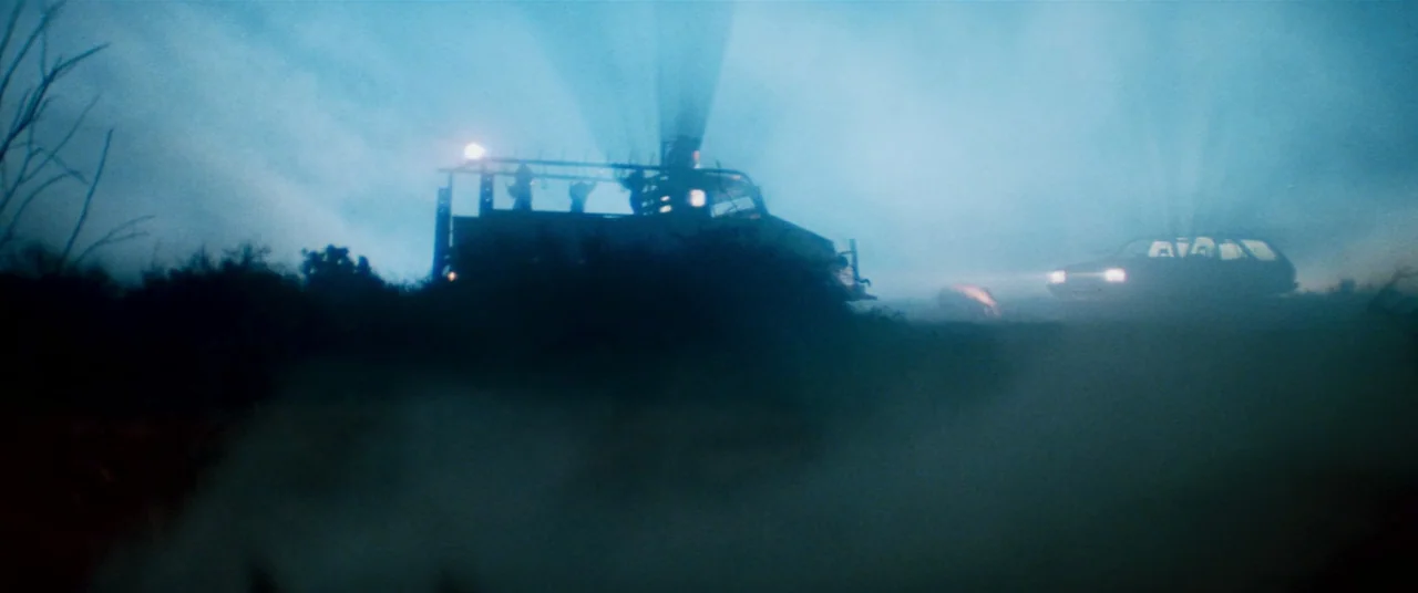 Razorback (1984) movie still: wide — A dark, fog-filled landscape at night features two vehicles with active light beams. The silhouettes of the…