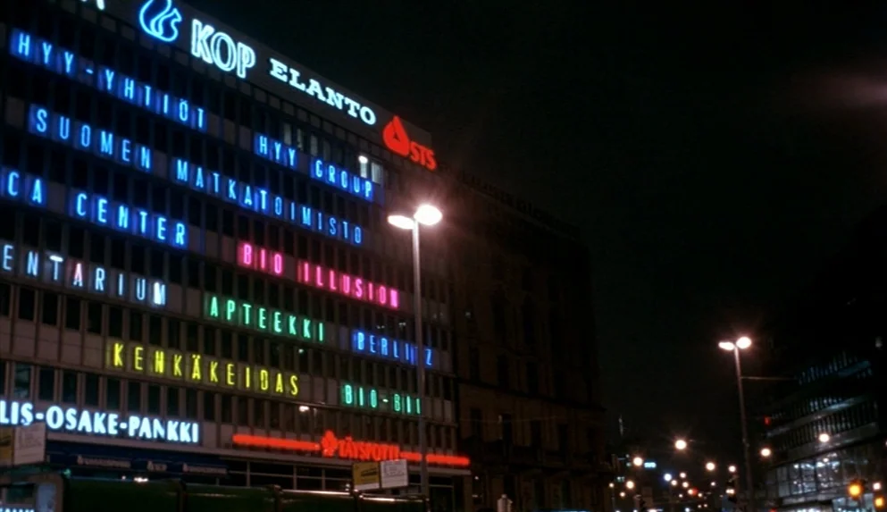 Night on Earth (1991) movie still: establishing — A dark city street building facade is covered in numerous glowing neon business signs. The facade…