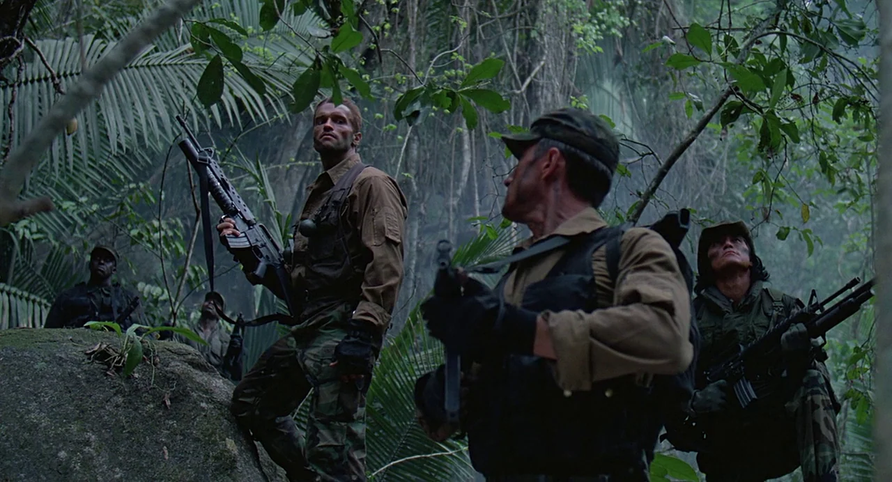 Predator (1987) movie still: wide — A dense, humid jungle environment filled with thick vegetation and tall trees. Four soldiers in tactical gear…