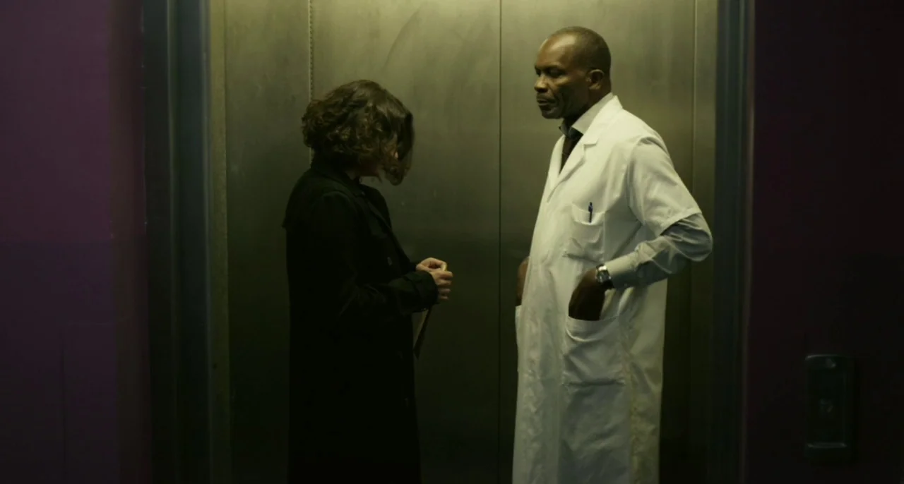 Bastards (2013) movie still: medium — Two people stand inside a metal-walled lift with a simple push-button interface panel. A doctor in a white…