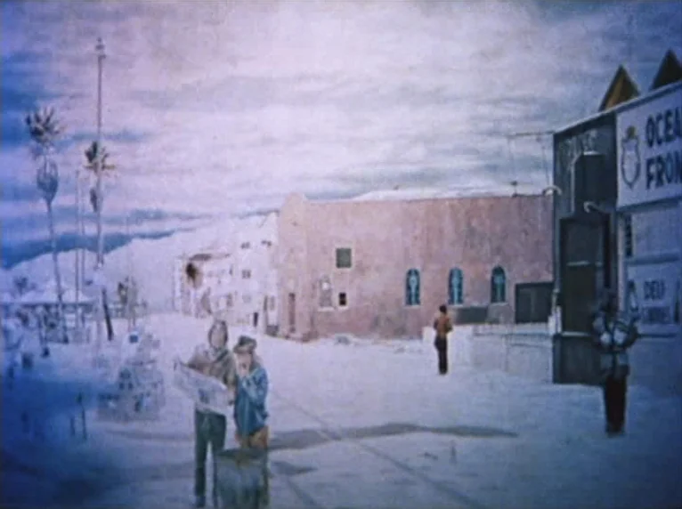 Mur Murs (1981) movie still: wide — A seaside boardwalk area features stucco buildings, palm trees, and signage under a cloudy sky. Two men stand in…