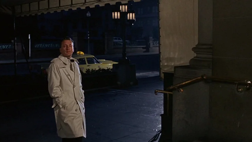Petulia (1968) movie still: fullbody — An exterior hotel entrance at night with stone pillars and a dark canopy overhang. A man in a trench coat…