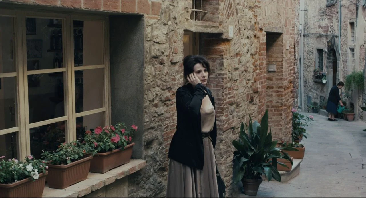 Certified Copy (2010) movie still: fullbody — A narrow alleyway with textured brick walls and potted flowers along the base. A woman in a dark…
