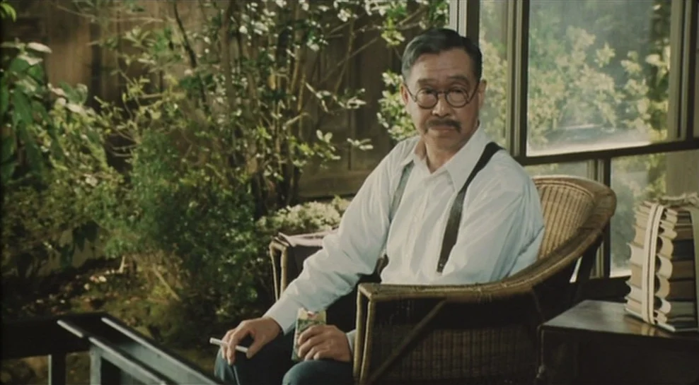 Madadayo (1993) movie still: medium — A glass-enclosed room with a wicker chair and a stack of tied books. An elderly man with a mustache sits while…