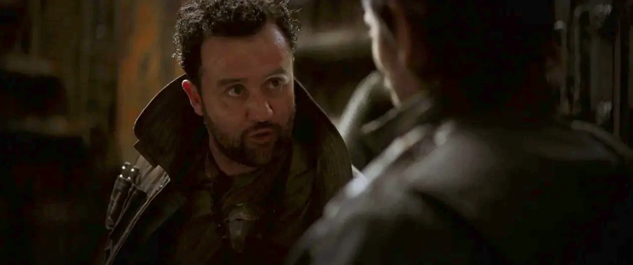 Rogue One: A Star Wars Story (2016) movie still: medium — Two individuals interact in a dimly lit, nondescript room with shadows. A bearded man with…