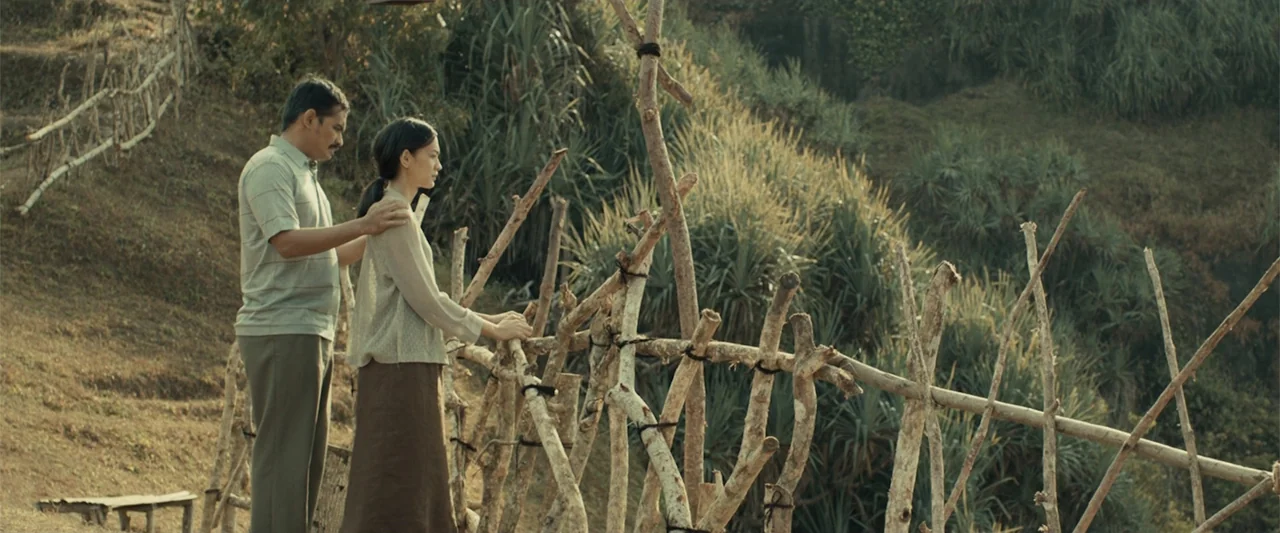 The Unholy (2022) movie still: fullbody — A rustic wooden stick fence borders a grassy hillside overlooking dense tropical vegetation. A man and a…