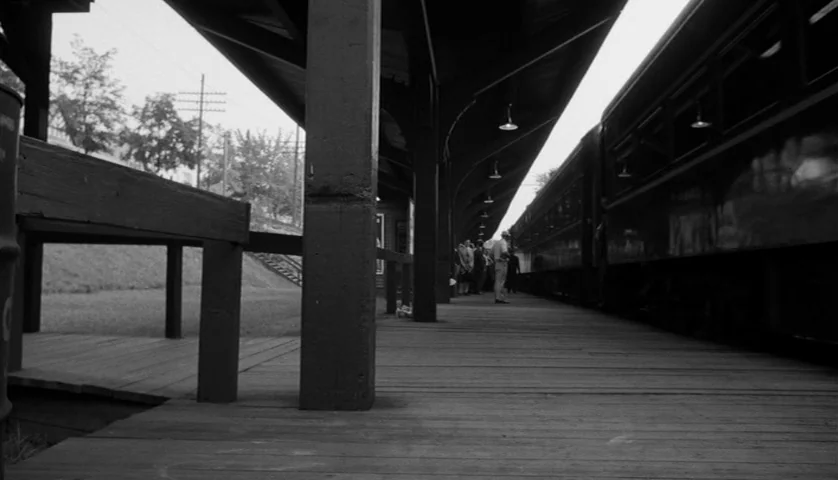 Seconds (1966) movie still: wide — A wooden train station platform features a long covered walkway with multiple overhead lamps. People stand near a…