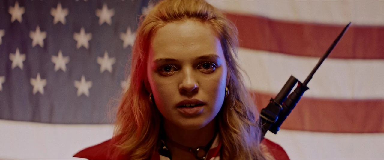 Assassination Nation (2018) movie still: closeup — A woman stands centered in front of a slightly out-of-focus United States flag. She holds a rifle…