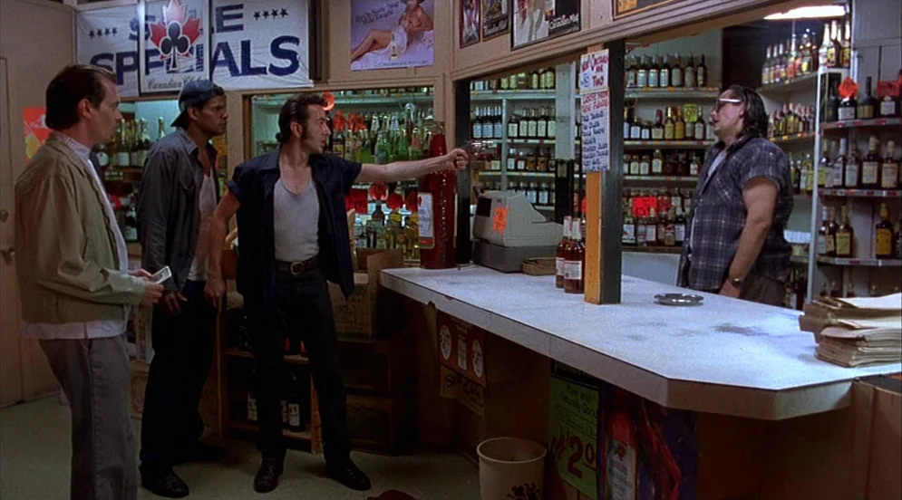 Mystery Train (1989) movie still: fullbody — A cluttered liquor store with wooden shelving packed with bottles and product posters. A man points a…