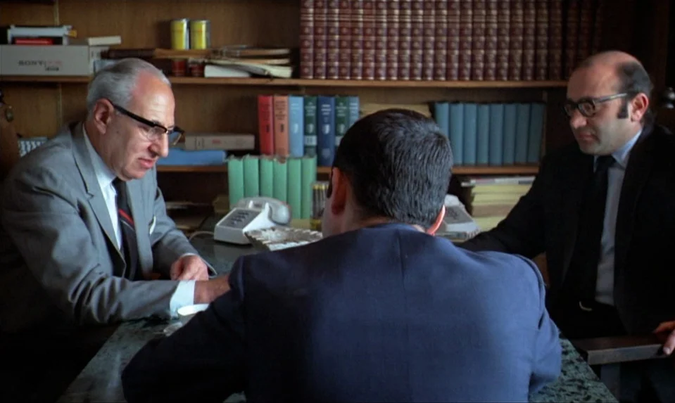 Lions Love (1969) movie still: medium — A wood-paneled room is lined with shelves filled with books and various boxes. Three men sit around a table…