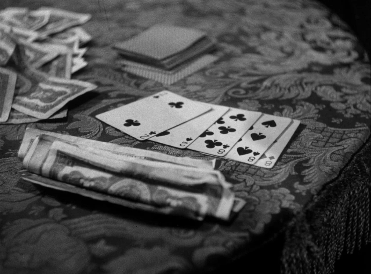 The Killers (1946) movie still: closeup — A table covered in a dark patterned fabric holds a game in progress. A deck of cards and a small stack of…