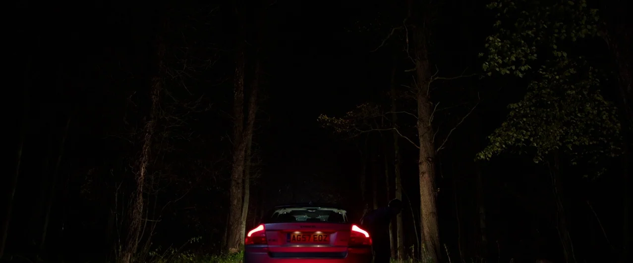 Ghost Stories (2017) movie still: wide — A silver passenger car is parked on a dark, tree-lined path at night. An obscured person stands next to the…