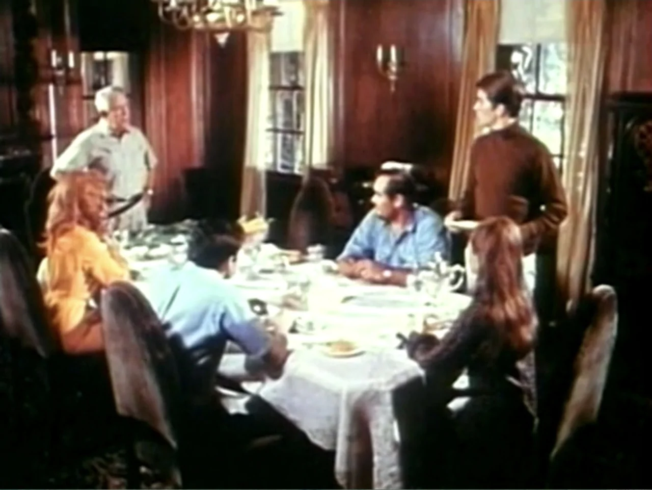 In the Year 2889 (1969) movie still: medium — A wood-paneled dining room contains a large table covered with a white tablecloth. Six individuals are…