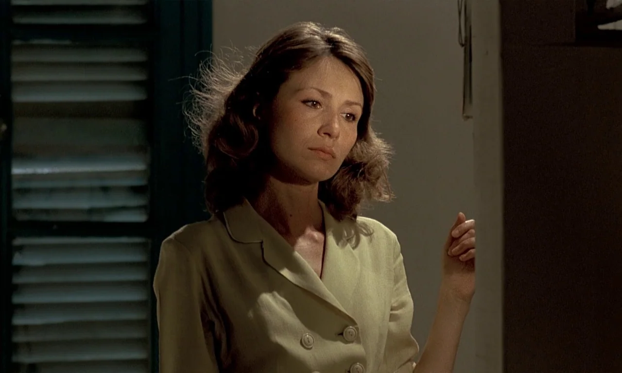 Coup De Torchon (1981) movie still: medium — A dimly lit room features a woman standing against a plain wall. A dark window shutter is visible in…