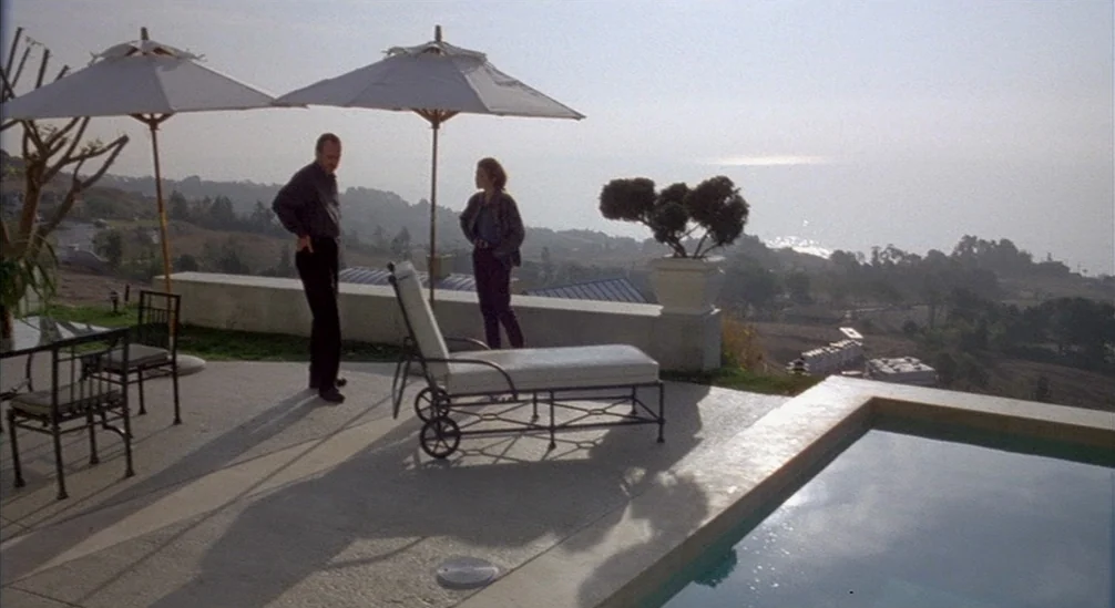 New Nightmare (1994) movie still: wide — A concrete patio beside a swimming pool overlooks a coastal hillside during the day. A man and a woman…