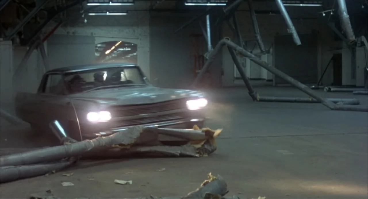 Cooley High (1975) movie still: wide — A spacious, dimly lit concrete industrial garage contains dangling metal ventilation ducts and debris. A grey…