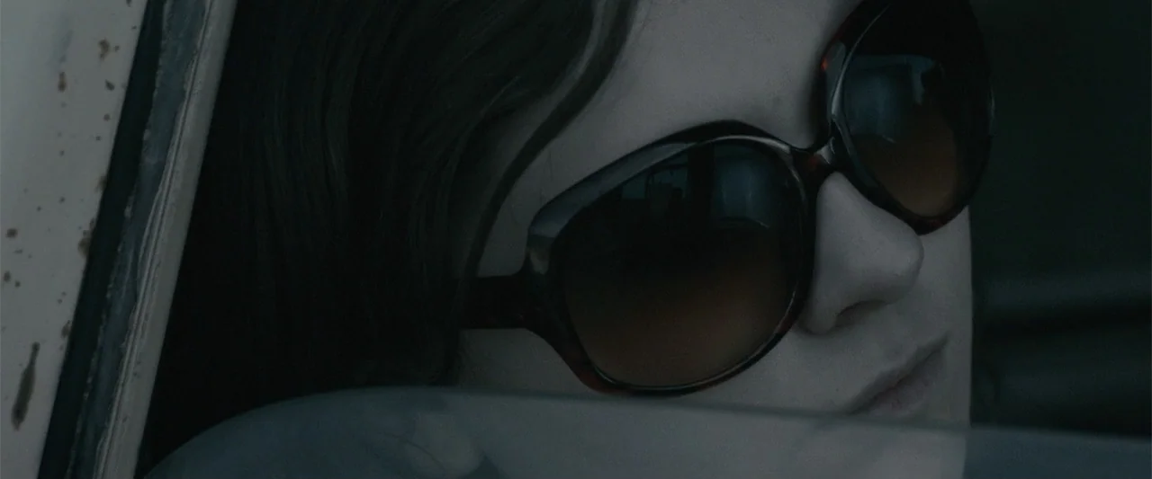 Maggie (2015) movie still: closeup — A dark car interior with a person leaning against the side window. A young woman with dark hair wears large…
