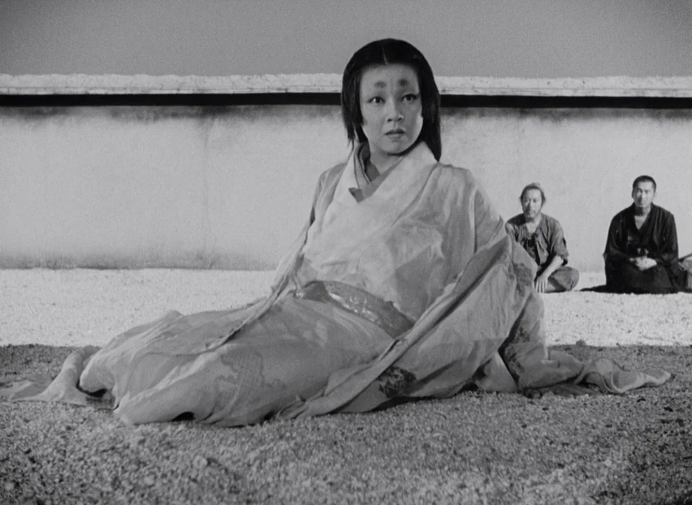 Rashomon (1950) movie still: fullbody — A woman in historical Japanese attire lies on a bed of light gravel. Two men sit cross-legged behind her in…