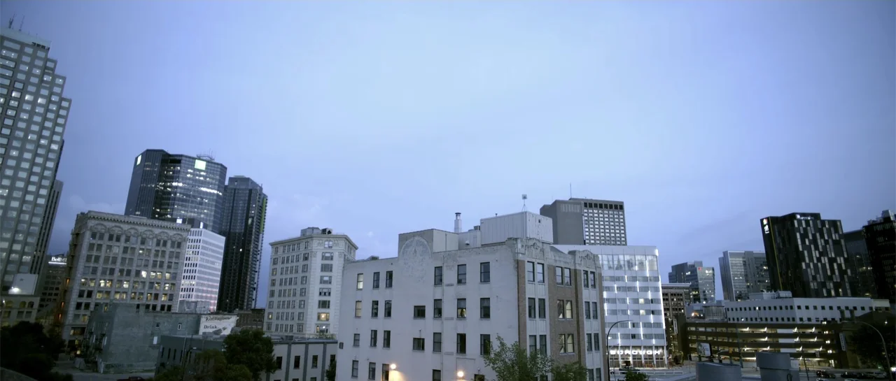 Elevator Game (2023) movie still: establishing — An urban skyline features a mix of older mid-rise buildings and modern high-rises. The city is…