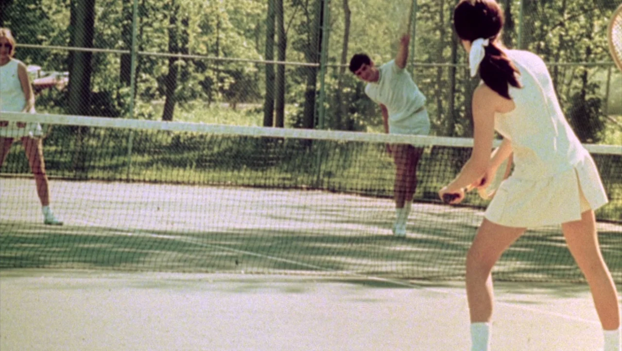 Shivers (1975) movie still: fullbody — An outdoor tennis court surrounded by tall green trees and a chain-link fence. Three players in white tennis…
