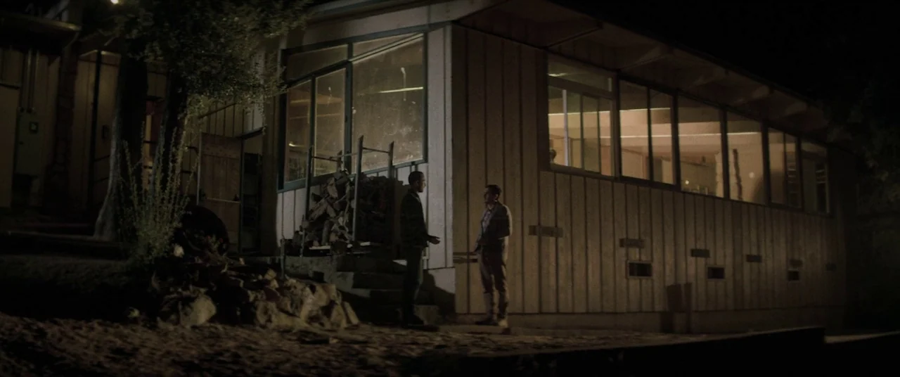 The Endless (2017) movie still: wide — A modern architectural house with vertical wooden siding stands in darkness at night. Two men stand together…