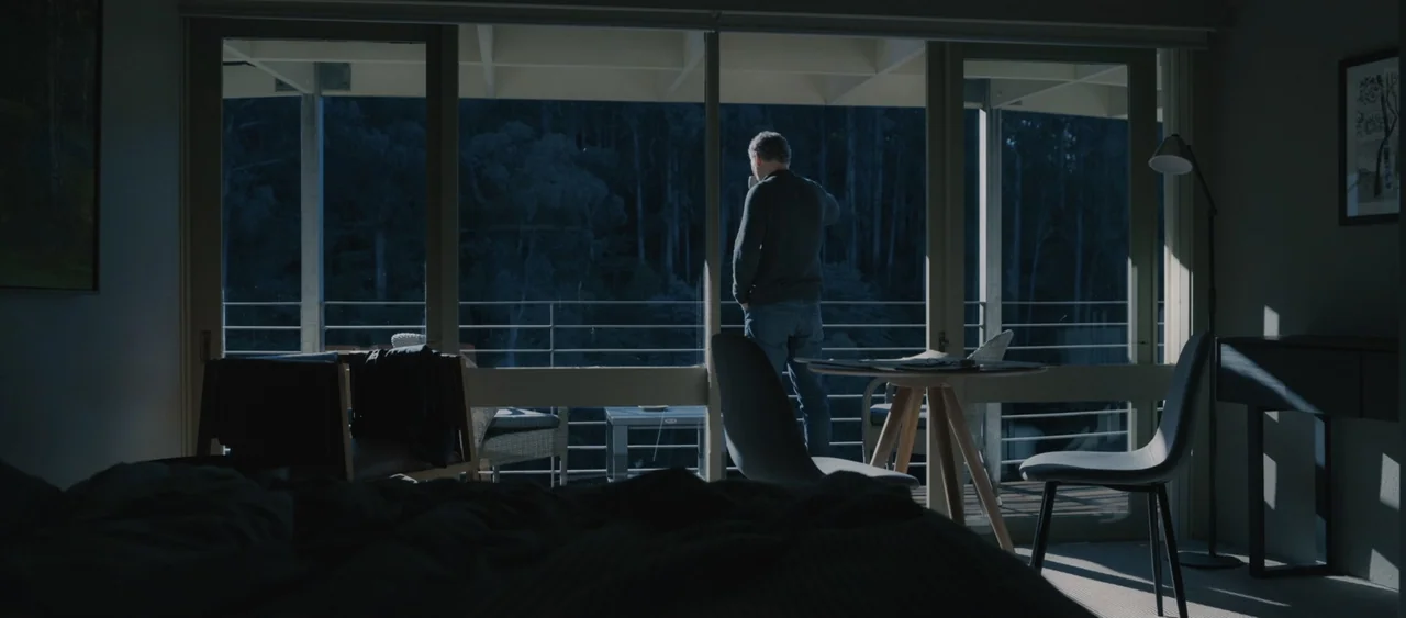 Force of Nature: The Dry 2 (2024) movie still: fullbody — A modern interior space features floor-to-ceiling windows overlooking a dense, dark forest…