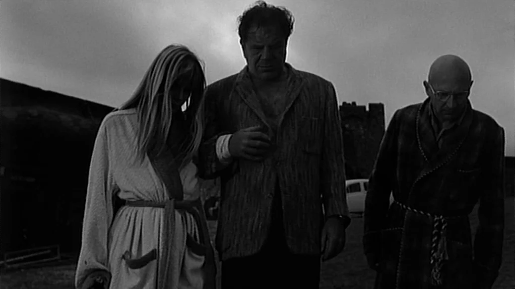 Cul-de-sac (1966) movie still: fullbody — A desolate field under a heavy overcast sky features a stone fortress in distance. Two men and a woman…