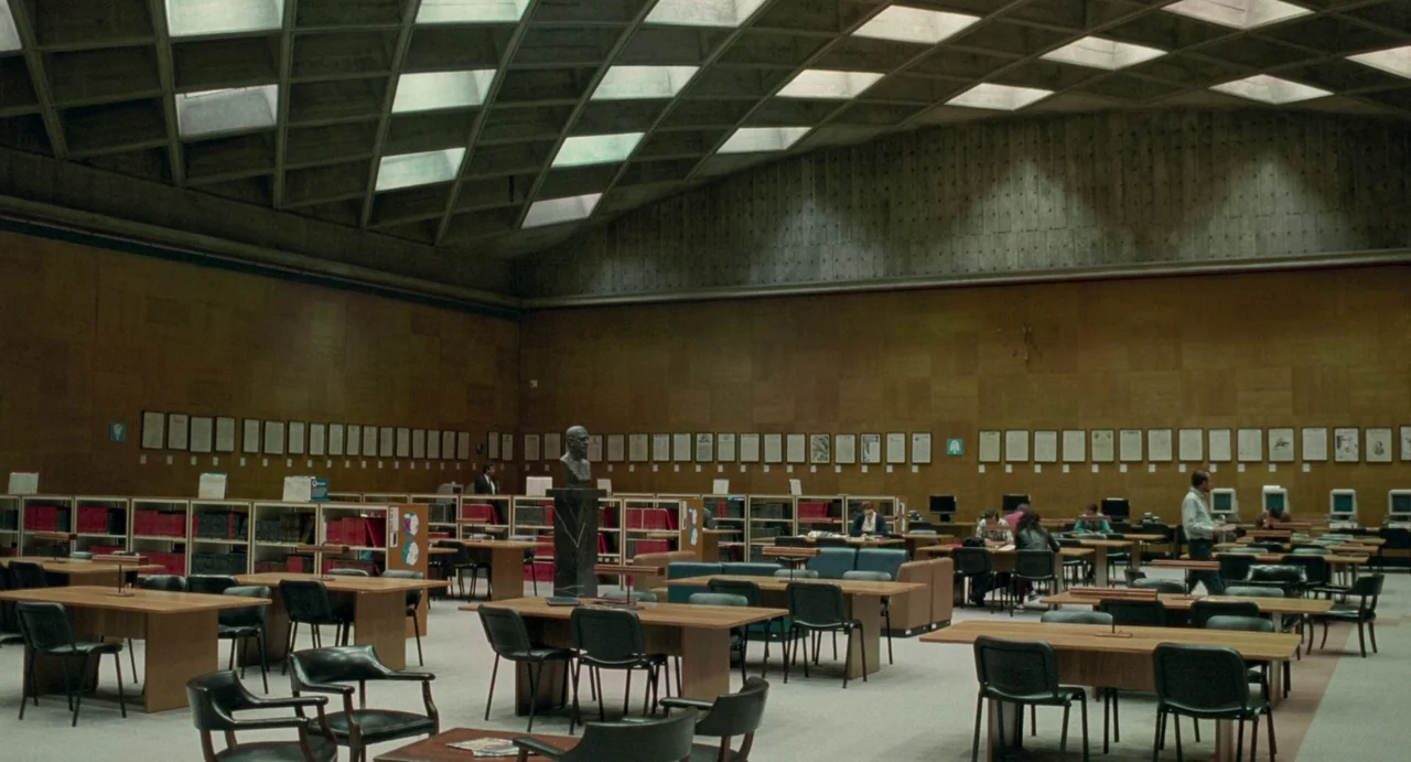 Memoria (2021) movie still: wide — A spacious library interior features a distinctive coffered ceiling with inset glass skylights. Several people…