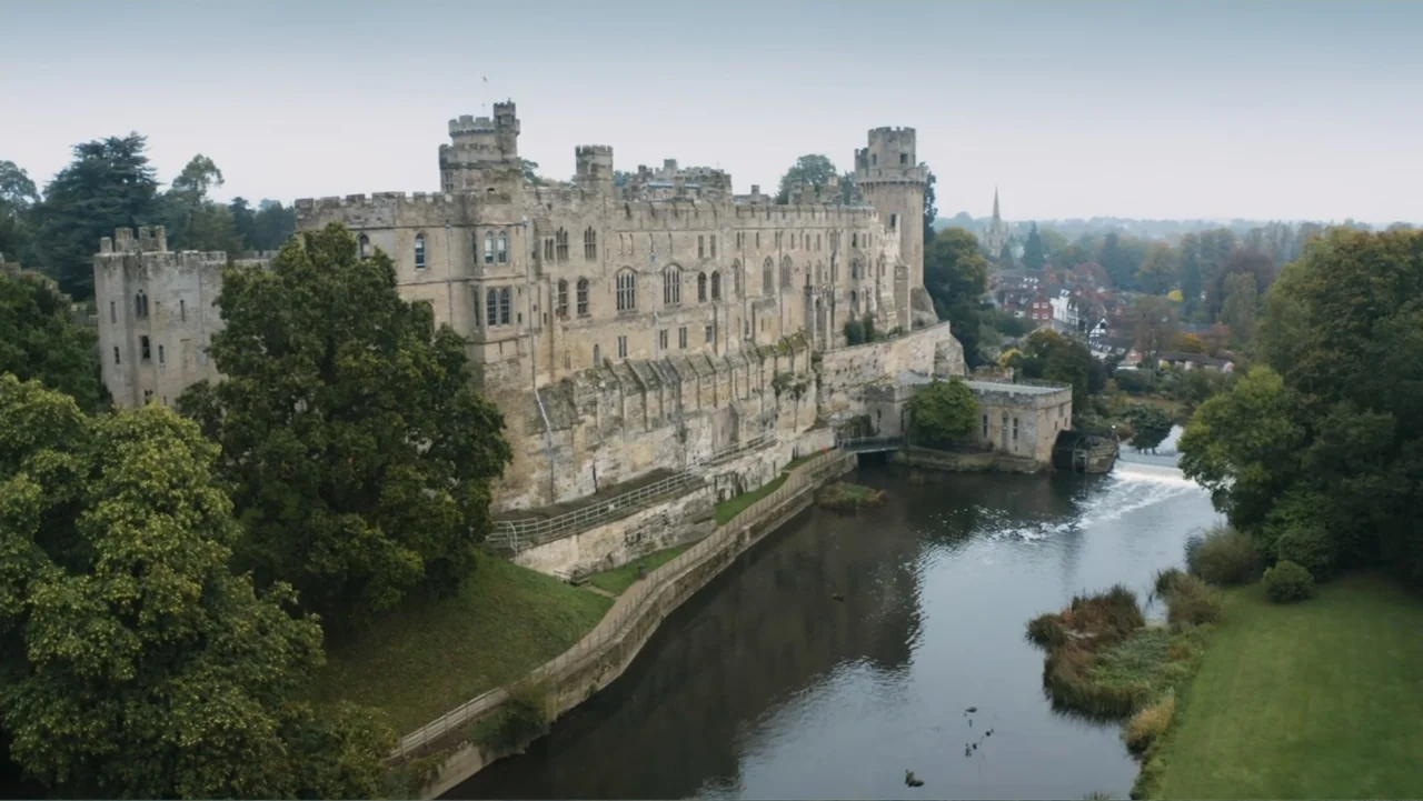 Fantastic Beasts: A Natural History (2022) movie still: establishing — A stone castle complex rises above a river with lush green trees lining the…