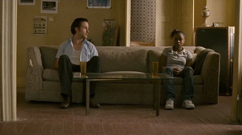 Half Nelson (2006) movie still: wide — A spare, beige-walled space features a sofa against a wall with framed pictures. A man and a woman sit at…