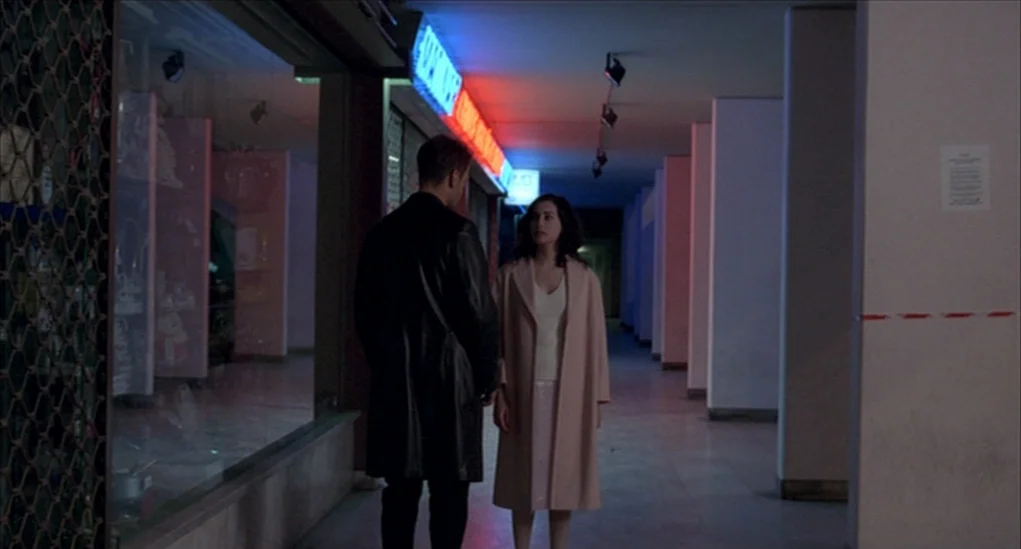 Anatomy of Hell (2004) movie still: fullbody — A quiet shopping mall corridor features large glass store fronts and repetitive concrete pillars. A…