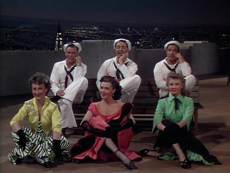 On The Town (1949) movie still: wide — A group of six people sit on a rooftop overlooking a distant illuminated city. Three sailors sit on a bench…