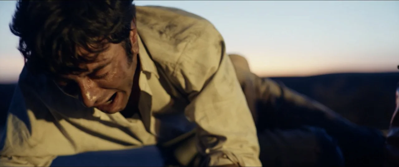 Hear Me: Our Summer (2024) movie still: medium — An open desert landscape at golden hour with distant flat horizon lines. A man in a dirt-stained…